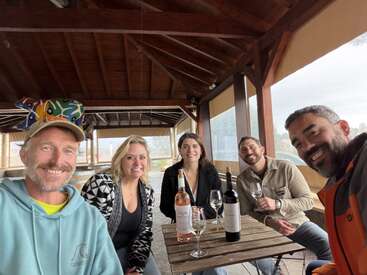 Five friends sit around a wooden table in an outdoor covered patio, smiling and enjoying wine together. Two wine bottles and glasses are on the table.