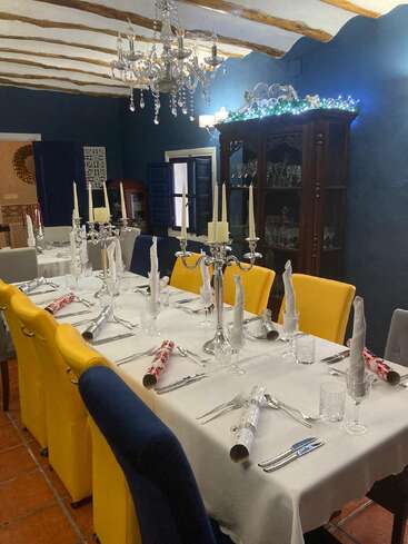 Uma sala de jantar formal preparada para uma comemoração, com cadeiras amarelas e azuis, castiçais elegantes, toalha de mesa branca, biscoitos festivos e um lustre cintilante.