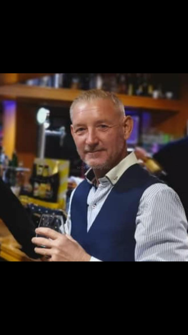 Um homem com cabelo curto e barba aparada está sorrindo, segurando uma bebida, vestido elegantemente com um colete azul, em um bar animado.