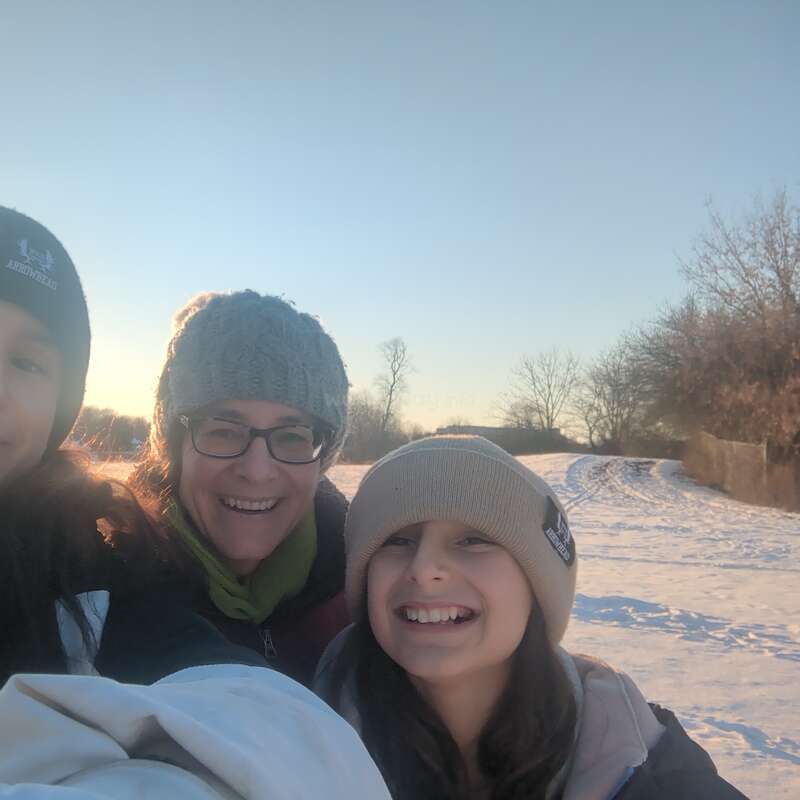 Drei Menschen, dick eingemummt in Winterkleidung und Mützen, lächeln herzlich für ein Selfie in einer verschneiten Landschaft bei Sonnenuntergang, umgeben von Bäumen und einem strahlend blauen Himmel.
