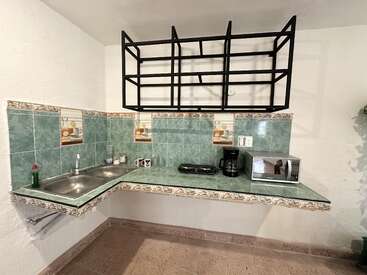This small kitchen features green-tiled counters and backsplash, a stainless steel sink, electric stove, coffee maker, microwave, mugs, and a modern black open shelf.