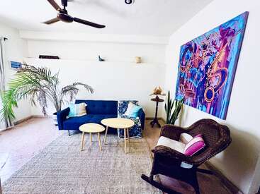 This cozy living room features a blue couch, wicker rocking chair, two wooden tables, indoor plants, abstract colorful wall art, ceiling fan, and neutral decor accents.