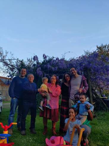 The image depicts a family of eight posing for a photo in a backyard, surrounded by a vibrant purple flowering bush and lush green grass.