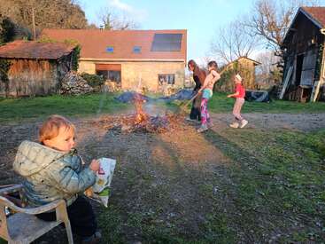 The image depicts a serene backyard scene with a family gathered around a small bonfire, surrounded by a rustic house and lush greenery on a sunny day.