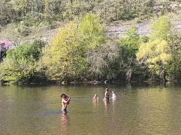 The image depicts four individuals wading in a river, surrounded by lush greenery and trees, with a hillside covered in vegetation in the background.