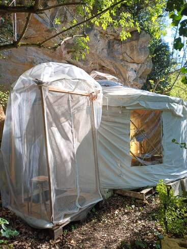The image depicts a small, makeshift structure with a white tarp covering and a wooden frame, situated in front of a large rock formation in a wooded area.