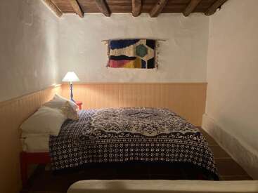 Este acogedor dormitorio rústico cuenta con un techo de madera, paredes blancas, una colcha estampada, un tapiz de colores, una lámpara de mesilla y un estilo decorativo sereno y minimalista.
