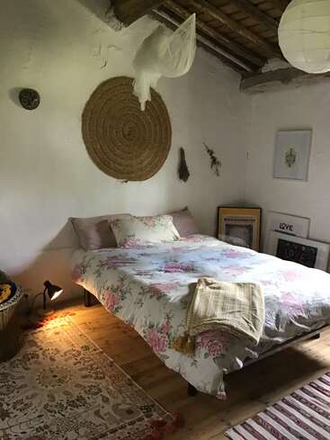 Un acogedor dormitorio rústico con una colcha de flores, adornos de pared tejidos, techo de madera, una lámpara colgante, alfombras estampadas, cuadros enmarcados y flores secas, que crean un ambiente tranquilo.