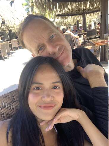 Una mujer joven y un hombre mayor posan muy juntos, sonriendo afectuosamente. Están sentados al aire libre en un restaurante o cafetería tropical con techos de paja.