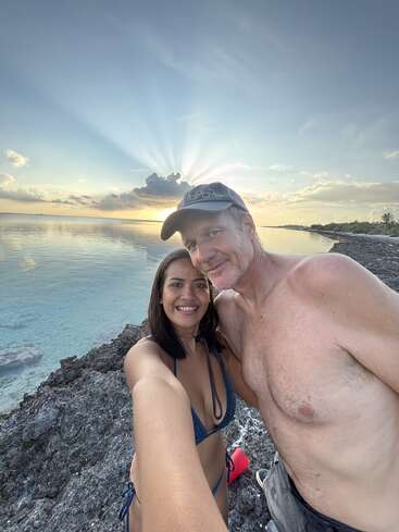 Una pareja sonriente se toma un selfie en la orilla rocosa al atardecer, con agua azul clara, un cielo espectacular, rayos de sol detrás de las nubes y árboles tropicales en la distancia.