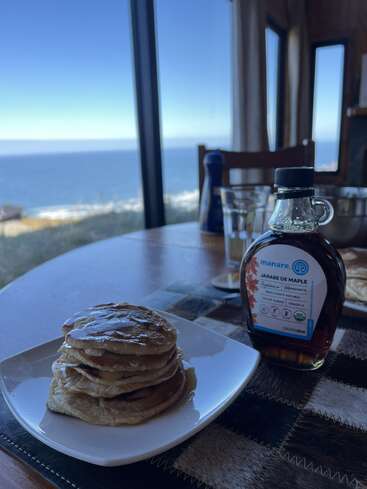 A stack of pancakes drizzled with maple syrup sits on a table by a window, overlooking a beautiful ocean view. Cozy and inviting breakfast scene.