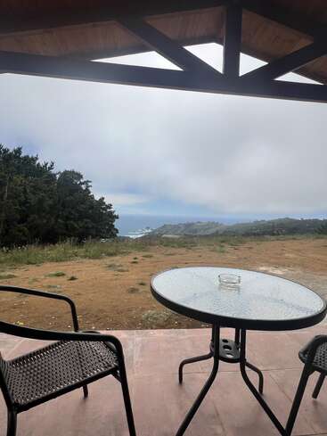A glass table and two chairs sit on a patio under a wooden roof, overlooking a grassy landscape with distant hills and a cloudy sky.