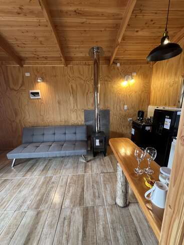 This cozy cabin interior features wooden walls and ceiling, a gray futon, wood stove, small kitchen area, breakfast bar, wine glasses, mugs, and rustic decor.