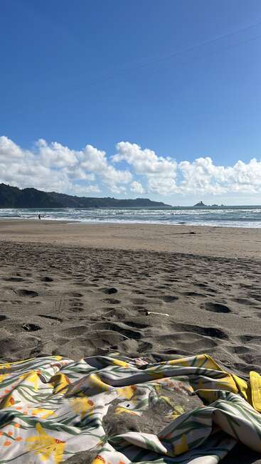 A bright, sunny beach scene with soft, golden sand, a floral-patterned towel, gentle waves, distant hills, scattered clouds, and a single person near the shoreline.