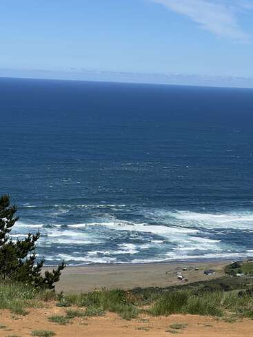 The image shows a breathtaking coastal view with deep blue ocean waves crashing onto a sandy beach, green grassy foreground, scattered beach houses, and a clear blue sky.