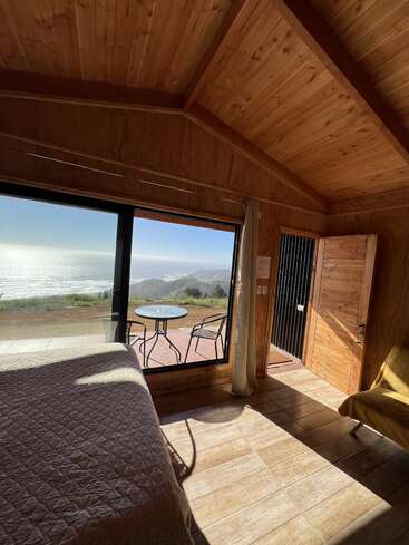 A cozy wooden cabin interior with sunlight streaming in, a bed, chair, open door, and large window offering a beautiful view of the ocean and coastline.