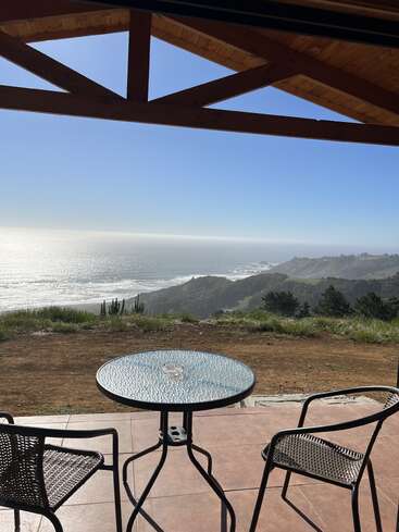 Two chairs and a glass table sit on a patio overlooking a beautiful coastal view, with blue sky, ocean waves, and green hills in the distance.