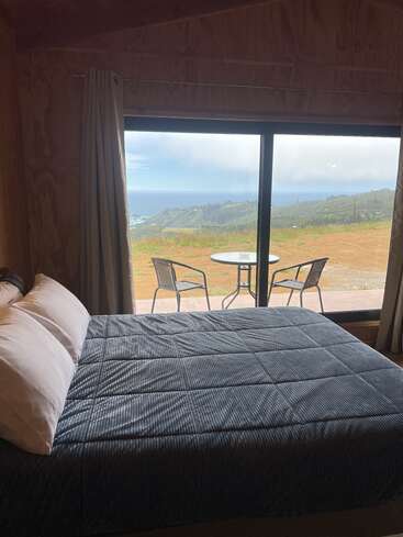 A cozy bedroom features a neatly made bed, large windows with curtains, and a breathtaking view of rolling hills, ocean, and a small outdoor seating area.