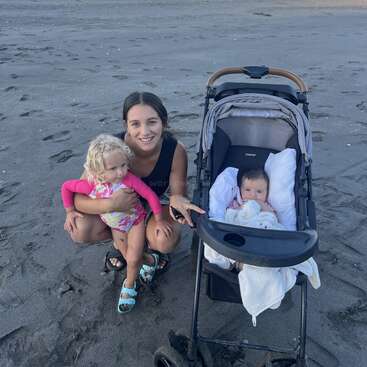 Une femme est agenouillée sur une plage de sable, serrant dans ses bras une jeune fille blonde. À côté d'elles, un bébé est assis dans une poussette. Des montagnes sont visibles à l'arrière-plan.