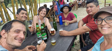 Six people sit around a wooden table outdoors, smiling and holding drinks. There are bottles and glasses of beer, lush greenery, and a relaxed, social atmosphere.
