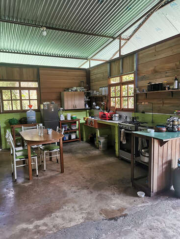 This image shows a rustic kitchen with wooden walls, metal roof, basic appliances, open shelving, a dining table with chairs, and concrete flooring, filled with natural light.