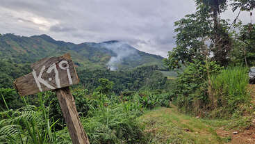 Un panneau en bois portant l'inscription « K:19 » se dresse au bord d'un chemin de terre, au milieu de collines verdoyantes. De la brume et de la fumée lointaine s'élèvent au milieu d'une végétation tropicale dense et des montagnes.