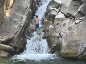 Uma pessoa sobe por um estreito desfiladeiro rochoso, cercado por penhascos escarpados, com água caindo em cascata por baixo deles, criando uma pequena cachoeira em um ambiente natural.