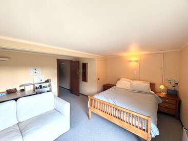 A cozy bedroom with a wooden double bed, nightstands, a comfortable white sofa, desk area with shelves, cream walls, soft lighting, and minimalistic decor.