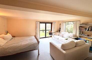This cozy, light-filled bedroom features a bed, two white sofas, bookshelves, a guitar, large windows with curtains, and a glass door opening outside.