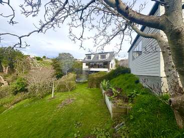 A modern two-story house with large windows sits on a lush, green lawn surrounded by trees, shrubs, and garden beds. Peaceful, natural outdoor setting.