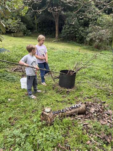 Two children standing on green grass, holding long sticks, beside a metal barrel filled with branches. Nearby is a tree stump with rocks lined on top.