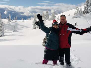 A joyful couple poses together in deep, fresh snow, surrounded by snowy trees and mountains under a bright sky, celebrating a beautiful winter day outdoors.