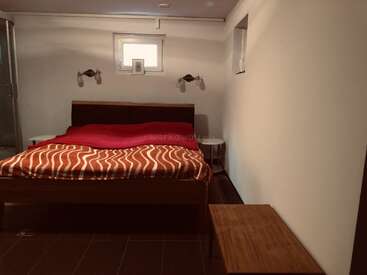 Un dormitorio minimalista cuenta con una cama de matrimonio con una manta ondulada roja y naranja, dos mesitas auxiliares, lámparas de pared, un banco y dos pequeñas ventanas.