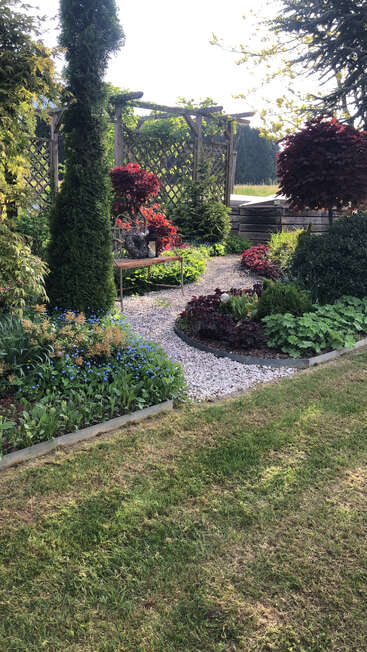 Eine ruhige Gartenszene mit saftig grünem Gras, blühenden Pflanzen, einem Kiesweg, einer Holzlaube, einer dekorativen Bank und schön gepflegten Sträuchern, die im Sonnenlicht liegen.