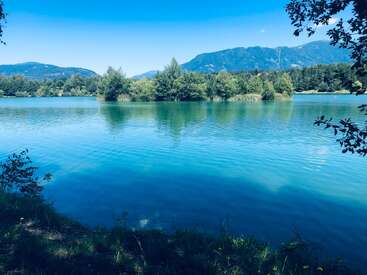 Ein ruhiger See mit klarem, blauem Wasser, umgeben von üppig grünen Bäumen und fernen Bergen unter einem hellen, sonnigen Himmel, der eine friedliche Naturszene schafft.