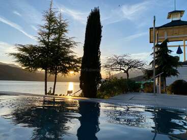 Eine ruhige Seeszene bei Sonnenuntergang mit hohen Bäumen, einem ruhigen Pool, in dem sich der Himmel spiegelt, ruhigem Wasser, entfernten Hügeln und einem lichtdurchfluteten Pavillon.