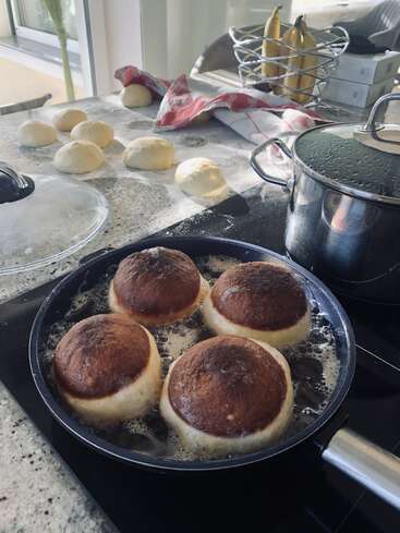 Vier Krapfen werden in einer Pfanne auf dem Herd gebraten, während mehrere Teigkugeln auf einer bemehlten Arbeitsplatte liegen. Küchengeräte und Bananen liegen in der Nähe.