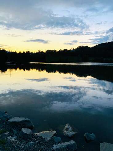 Ein ruhiger Blick auf den See in der Abenddämmerung, Felsen im Vordergrund, ruhiges Wasser, in dem sich der gelbe und blaue Himmel spiegelt, Silhouetten von Bäumen und in der Ferne sichtbare Hügel.