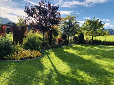 Ein üppiger, grüner Rasen, umgeben von bunten Bäumen, Büschen und Blumenbeeten. Das Sonnenlicht wirft lange Schatten, die einen friedlichen, gut gepflegten Garten unter einem hellen Himmel hervorheben.