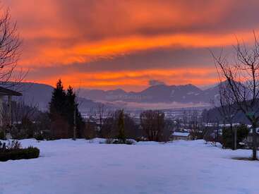 Eine atemberaubende Schneelandschaft bei Sonnenuntergang, leuchtend orangefarbene und rosafarbene Wolken am Himmel, Berge in der Ferne, kahle Bäume und eine ruhige Winteratmosphäre. Friedlich und heiter.