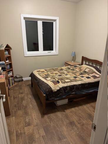 Este acogedor dormitorio cuenta con una cama individual con colcha de retazos, suelo de madera, estantería llena de libros, mesilla de noche con lámpara y una ventana.