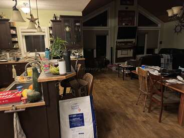 Acogedora cocina y sala de estar con suelos de madera, armarios oscuros, plantas y encimeras desordenadas. La mesa de comedor y la zona de TV contribuyen a crear un ambiente hogareño.
