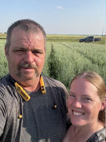 Un hombre y una mujer sonríen para hacerse un selfie en un campo soleado, con verdes cultivos a sus espaldas y una camioneta aparcada al fondo bajo un cielo despejado.