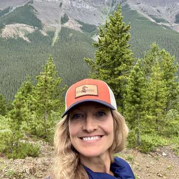 A smiling woman wearing an orange hat stands outdoors, surrounded by lush green pine trees with a backdrop of scenic mountains and a dense forest.