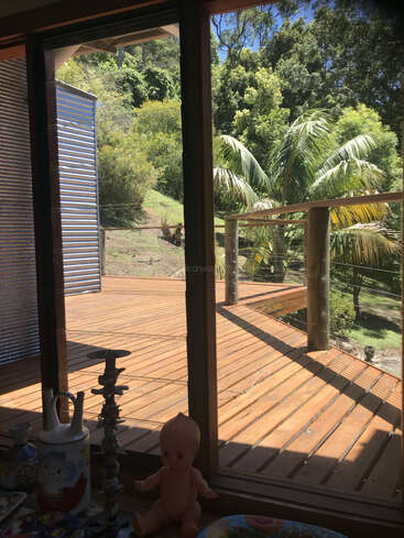 A vista através de uma porta de vidro mostra um deck de madeira, palmeiras verdes exuberantes do lado de fora, utensílios domésticos espalhados e uma boneca sentada perto da janela do lado de dentro.