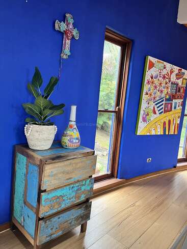 Uma parede azul vibrante apresenta uma cruz colorida, uma gaveta de madeira rústica, um vaso de plantas, um vaso decorativo, uma janela grande e uma pintura caprichosa e brilhante com flores e casas.