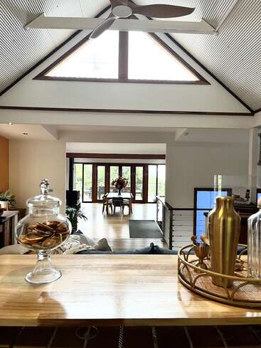 Uma moderna sala de estar em plano aberto com tetos altos abobadados, grandes janelas triangulares, detalhes em madeira com estilo, uma área de jantar e itens decorativos elegantes em um balcão de madeira.