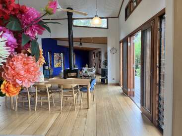 Uma sala de estar moderna e bem iluminada em plano aberto com piso de madeira, flores coloridas, uma mesa de jantar, fogão a lenha, parede com detalhes em azul e grandes janelas que dão para a vegetação.