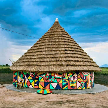 Uma cabana redonda tradicional com um grande telhado de palha, com as paredes decoradas com padrões geométricos coloridos e brilhantes, fica em um terreno aberto sob um céu azul impressionante.