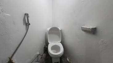 Esta imagem mostra um banheiro pequeno e simples com um vaso sanitário branco, uma tampa aberta, um pulverizador de bidê manual e um suporte de papel higiênico vazio em paredes brancas.